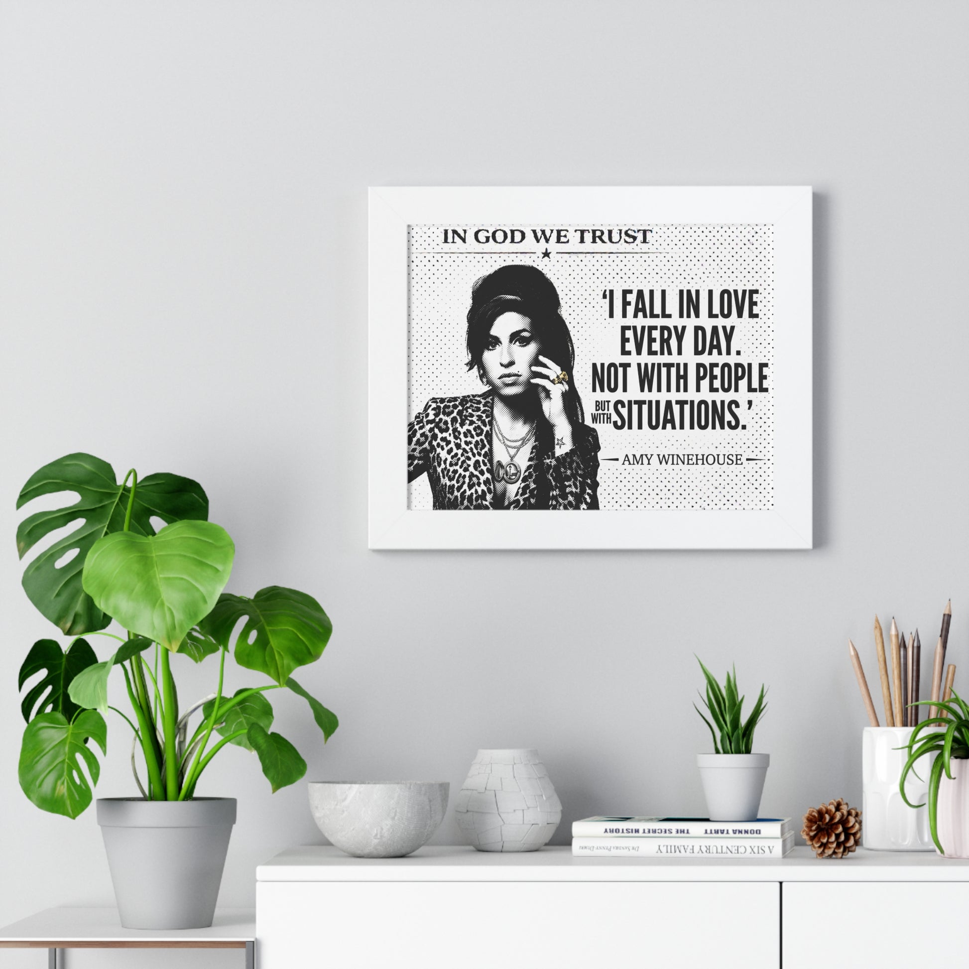 Framed artwork with a quote by Amy Winehouse on a wall above a desk with plants.