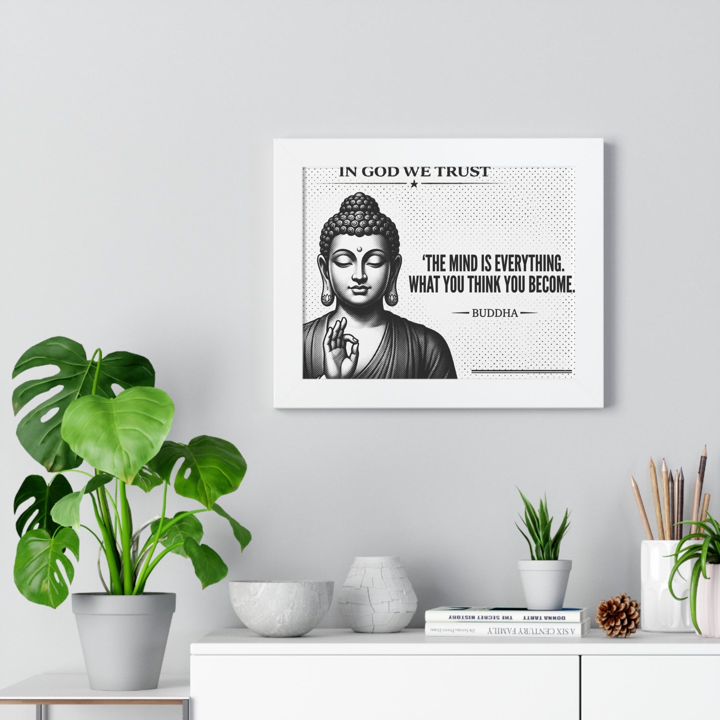 Framed artwork with Buddha image and quote on a wall with plants and decor below.