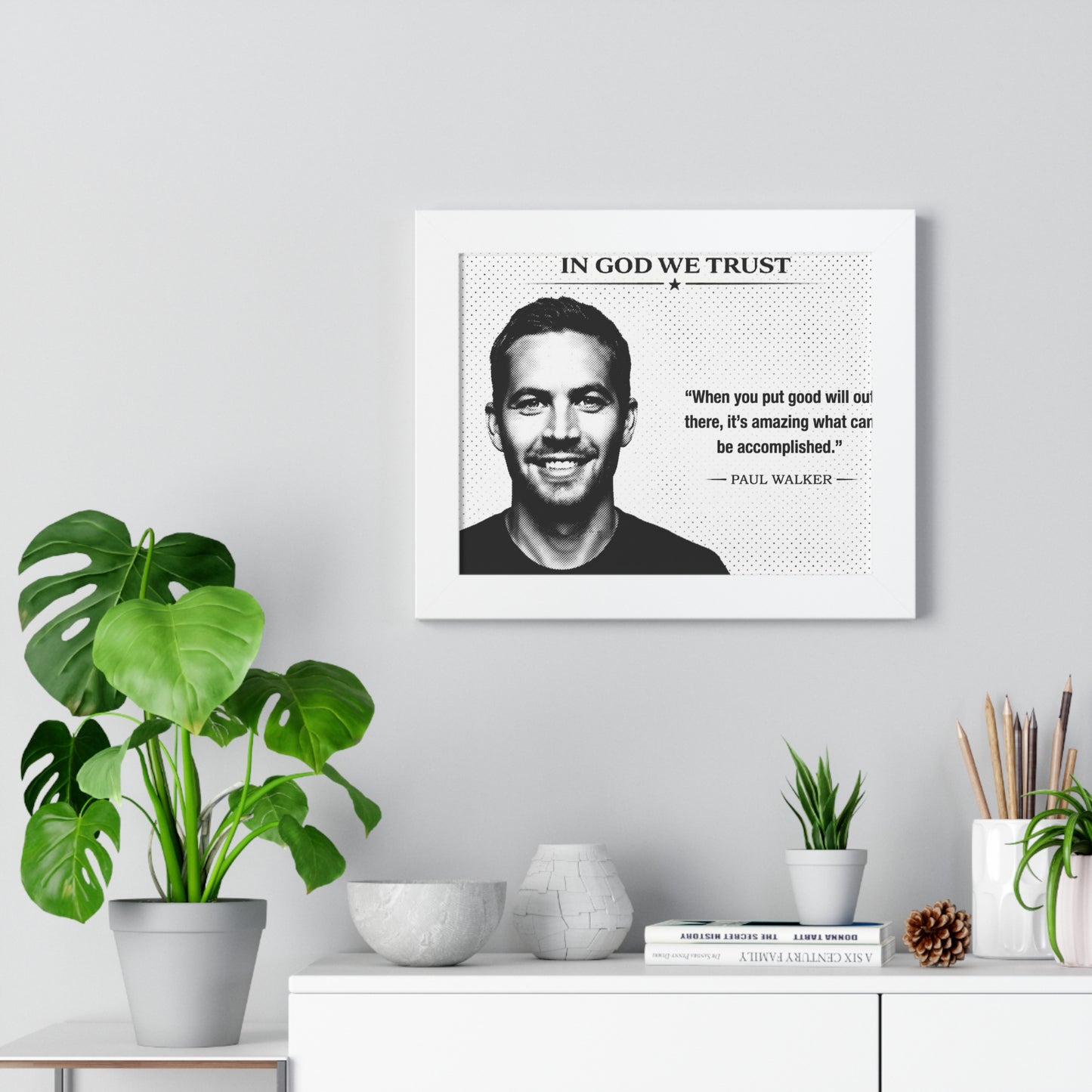 Framed portrait of a man with a quote on a wall above a white cabinet with decorative items.