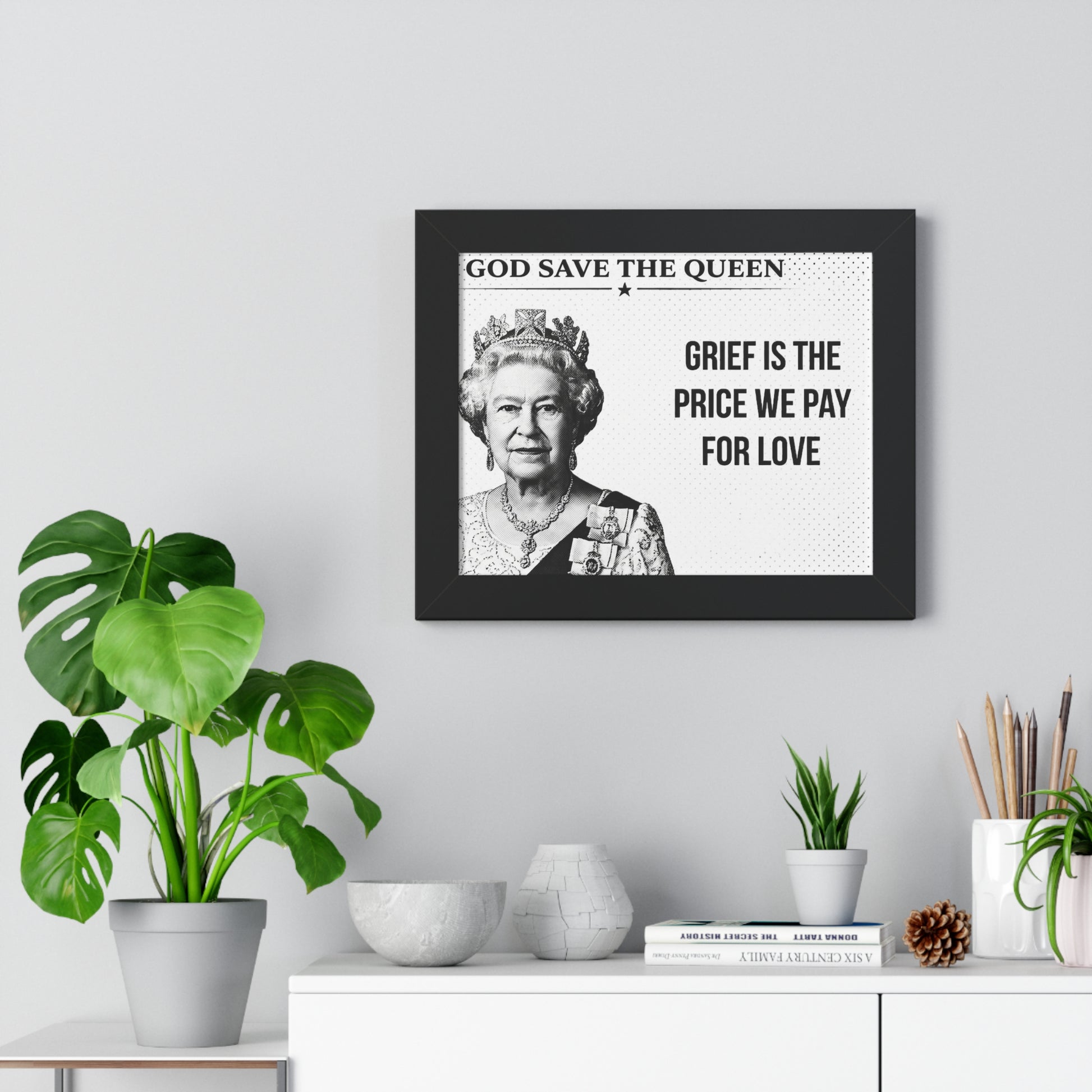 Framed portrait of a monarch with text above a desk with plants and decor.