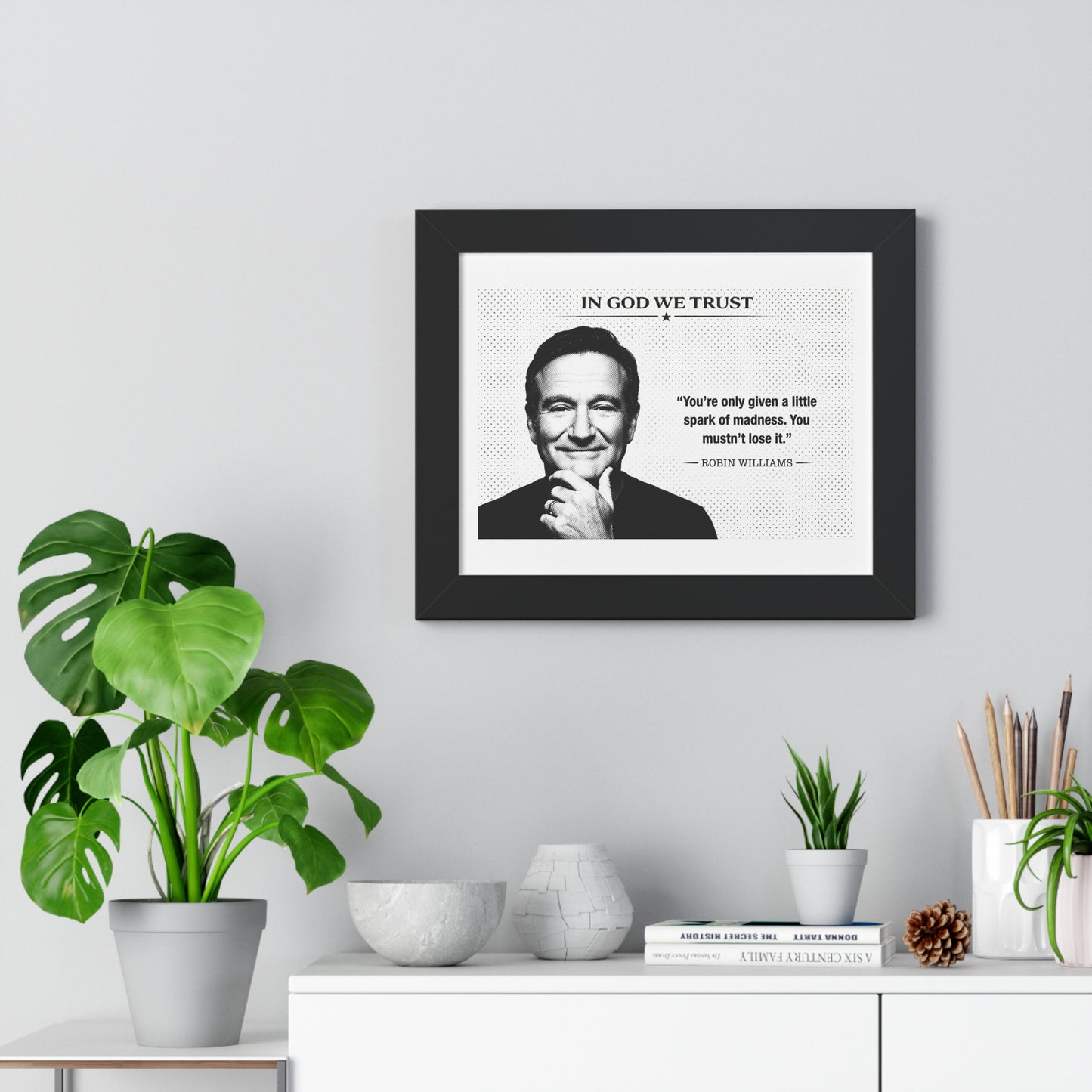 Framed print with a quote and portrait of a person on a wall above a white cabinet with decorative items.