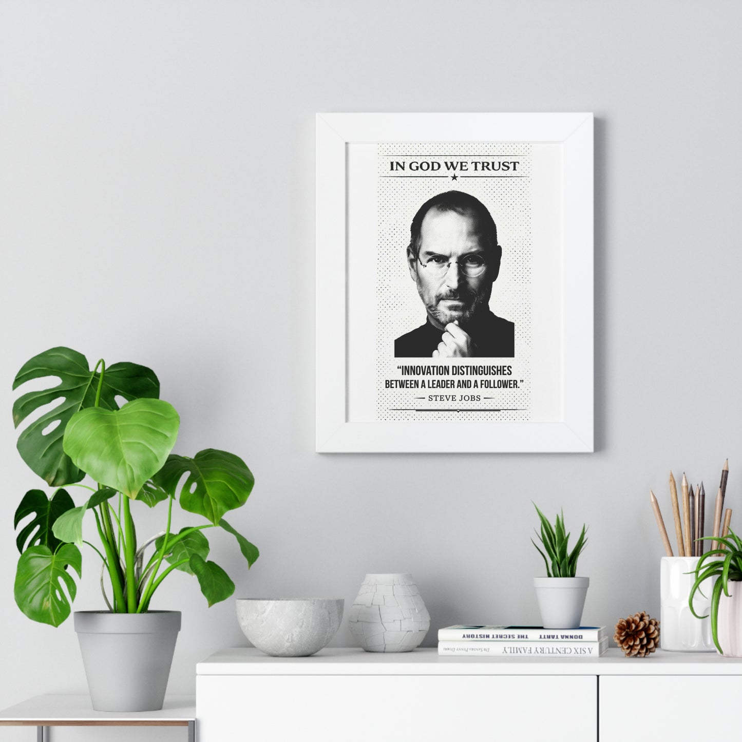 Framed poster with a portrait and quote on a wall above a desk with plants and decor.