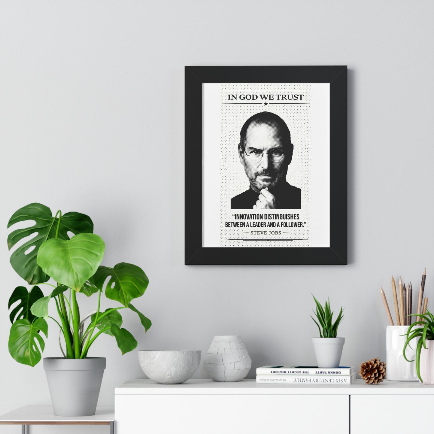 Framed poster with a portrait and quote of Steve Jobs on a wall above a white cabinet with decorative items.