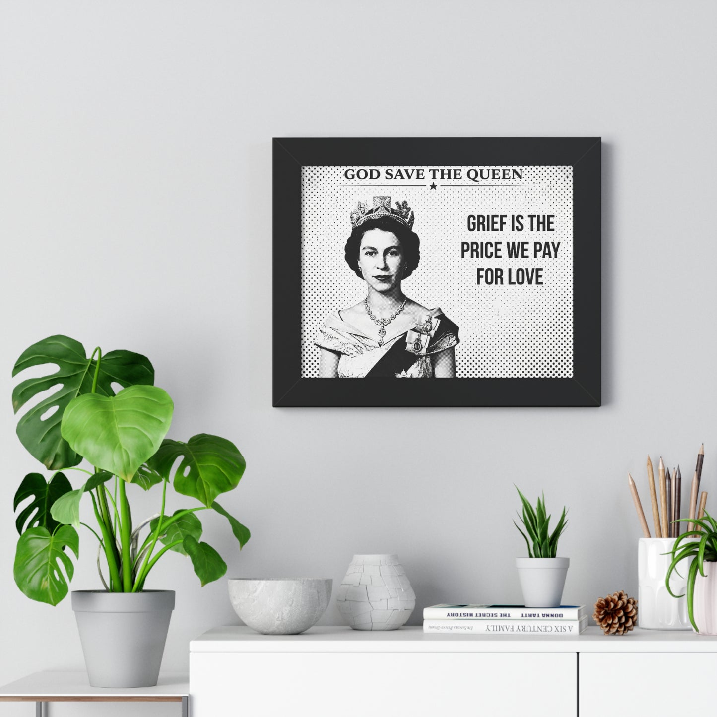 Framed artwork with a portrait of a queen and text on a wall above a white cabinet with decorative items.
